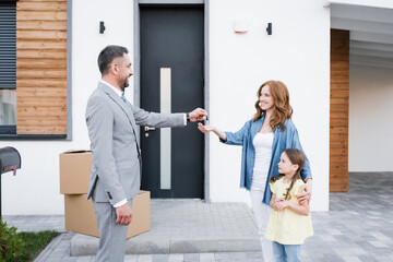 Happy broker giving key for woman with daughter near carton boxes on doorstep