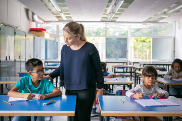 Serious school teacher watching pupils doing their task in class, sitting at desks and writing in...