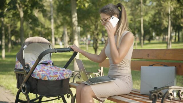 Confident busy Caucasian businesswoman in eyeglasses and elegant dress talking on the phone, typing on laptop keyboard, and rocking baby carriage. Young woman combining motherhood and business.