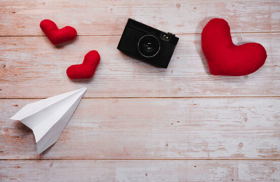 Wooden flat lay background for Valentines day holiday on February 14. White origami paper airplane, red stuffed plush toy hearts, and black old retro film camera.