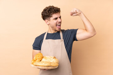Male baker holding a table with several breads isolated on beige background celebrating a victory