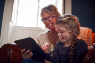 Granddaughter With Grandmother In Chair Looking At Digital Tablet At Home Together