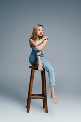 seductive topless woman in jeans looking away while sitting on high chair on grey