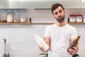 Portrait of thoughtful man standing with beer and milk in hands at home.