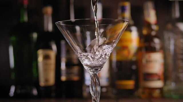 Close-up of a Martini being poured into a glass in a bar against a background of blurred bottles of various alcohol, slow motion.