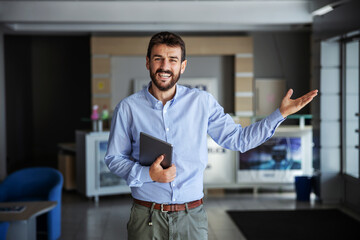 Smiling bearded CEO standing in lobby of shipping firm, holding tablet and gesturing delivery is on...