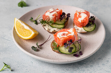 Mini open sandwiches with smoked salmon, cream cheese, cucumber and microgreen on rye bread, grey stone background. Appetizer with salmon and cheese cream sauce.