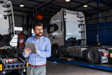 Young serious bearded CEO standing in garage of shipping firm and using tablet. In background are...