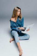 sensual barefoot woman in denim clothes, with naked shoulder, using laptop while sitting on grey