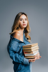 sensual woman in denim shirt, with naked shoulder, holding stack of books isolated on grey