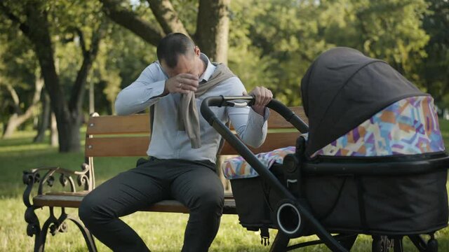 Yawning tired man rocking baby carriage and looking inside. Portrait of exhausted young Caucasian father sitting in sunny park with baby stroller. Fatherhood and parenthood concept.