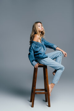 seductive young woman in denim shirt and jeans, with naked shoulder, sitting on high chair on grey