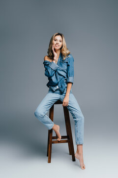 seductive, barefoot woman in stylish denim clothes sitting on high stool and touching face on grey