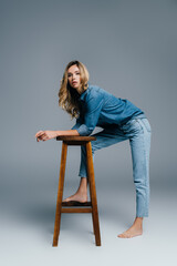 stylish barefoot woman in denim shirt and jeans leaning on high stool on grey