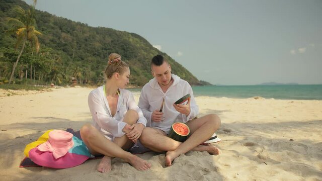 The cheerful love couple holding and eating slices of watermelon on tropical sand beach sea. Romantic lovers two people caucasian spend summer weekend in outdoor. Hat, backpack white shirt beachwear.