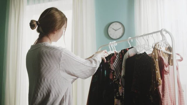 Girl in a white sweater and blue jeans stands in the middle of the room, trying on clothes and can not choose a dress