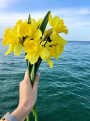 Foto op Canvas A girl holds a bouquet of yellow daffodils against the blue sea. Vertical photo. © Nataliia
