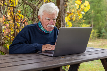 Old man working on laptop in his garden. A senior thoughtful man staring on the screen and making notes.