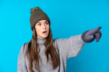 Teenager girl with winter hat over isolated blue background pointing away
