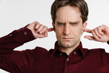 Handsome displeased young man plugging his ears and looking at camera