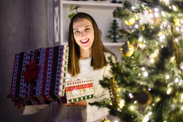 Attractive excitedly girl opening gift box sitting near festive Christmas tree. A girl in white sweater with a present box in hands looks at camera and smiles