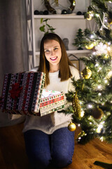 Attractive excitedly girl opening gift box sitting near festive Christmas tree. A girl in white sweater with a present box in hands looks at camera and smiles