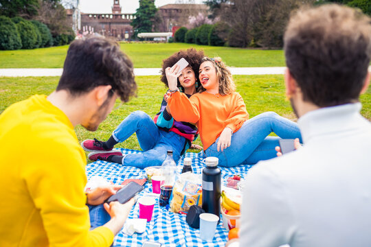 Four cheerful friends diverse multiethnic having fun doing pic nic in a park outdoors using smartphone