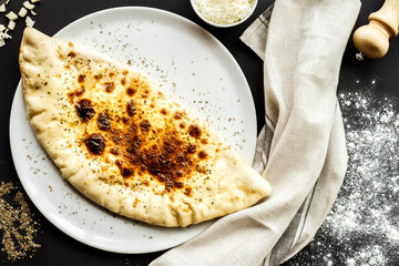 Crispy Calzone pizza with grated cheese and herbs on white plate, dark background.