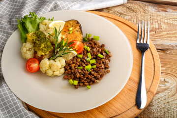 Baked fish with broccoli and lentils in a white plate with rosemary and lemon on a round stand next to a fork.