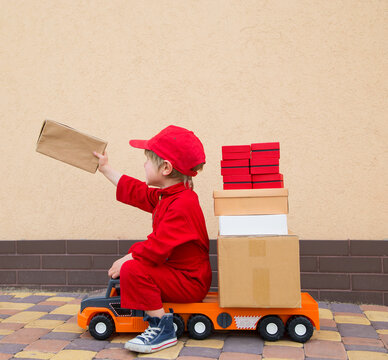 4-year-old boy in red overalls and a cap sits on big toy car - truck with many cardboard boxes on it. holds out the parcel with his hand. Parcel delivery, small postman, small truck driver. 