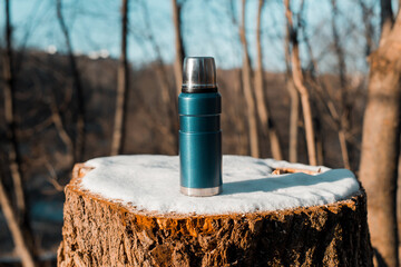 Thermos standing on tree stump covered with snow. Close-up of camping vacuum flask in the winter forest, sunny day.