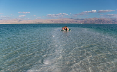 Obraz na płótnie Canvas Panoramic view of Dead sea coast near Ein Bokek resort, Israel. Young couple relaxing in the water of Dead Sea. Salt formations in the foreground and mountains in the background. Vacation in Israel.