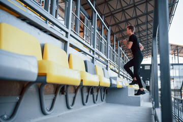 Running on the bleachers. Sportive young guy in black shirt and pants outdoors at daytime