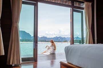 Travel Woman in White Dress Sitting Outdoor near Wooden House on Water. View on Tourist Woman Relaxing and Leisure from Raft House on Cheow Lan Lake in Thailand. Eco Tourism