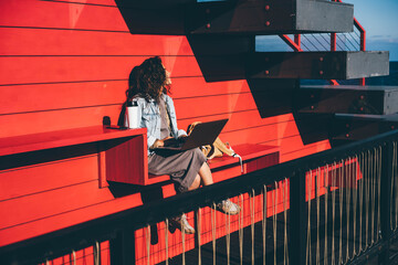 Pretty young woman in casual clothes with coffee cup and contemporary laptop sitting at the red bench on sunny autumn day.