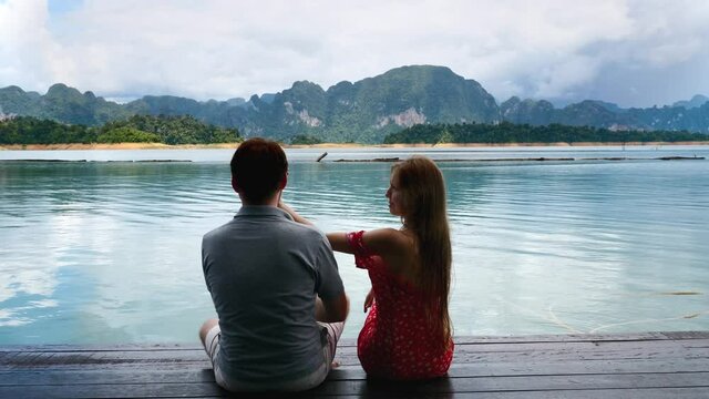 Rear View of Couple Sitting Together on Date on River Bridge, Man Flirting, Touching Woman Booty and Getting Funny Slap, Fun, Laughing and Kissing Together.Pair of Travel People at Lake with Mountains