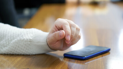A young man in a warm white sweater, sitting at a table, wants to use a blue smartphone. Pupil, student or participant in negotiations and meetings.
