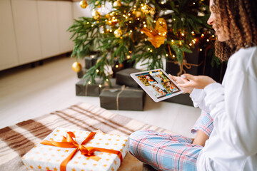Naklejka na ściany i meble Christmas online holiday. Happy woman having video call with their family or friends. Young woman uses a digital tablet near decorated festive tree at home. Virtual meeting. Covid-2019.