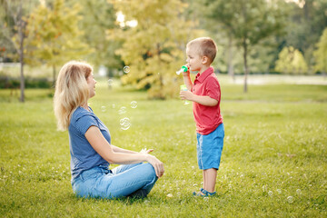 Mothers Day. Mother and boy toddler son blowing soap bubbles in park. Mom and child playing having fun together outdoors on summer day. Happy authentic family childhood lifestyle.