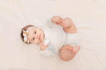 baby six months lies in a crib with a bottle and drinks water in a white bodysuit