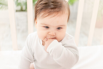 happy baby sitting in the crib in a white bodysuit and sucking his finger