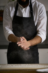 Naklejka premium The hands of the strong man knead the dough from which they will make bread, pasta, cake or pizza. In his kitchen he carries on the tradition of homemade pasta. A cloud of white flour flies like dust.