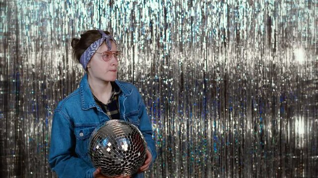 A young cocky girl from the 90s or 80s is spinning a disco ball in her hands on a shiny background. A beautiful model in a denim jacket tosses a ball and has fun.