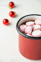 Top view of red mug with pink marshmallow and chocolate on white wooden table and defocused red christmas balls, vertical, with copy space