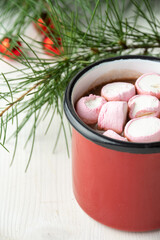 Top view of red mug with pink marshmallow and chocolate on white wooden table with green branches,...