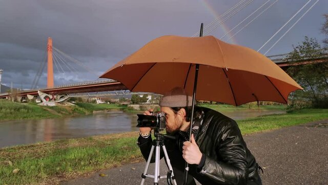 Caucasian photographer takes pictures under his umbrella during a rainy day, while smiling. Attractive man snapping some picture under the rain with rainbow behind him
