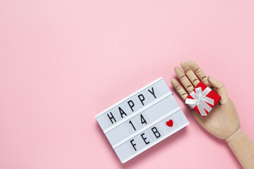 Trendy wooden human hand model holds small red gift box, light board with inscription Happy 14 feb on pastel pink background, copy space, flat lay. February 14th, Valentine's day celebration concept
