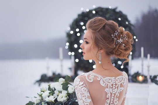 Beautiful girl in a lace wedding dress near the fireplace with Christmas decor on the background of snow-covered lakeand the night sky. Romantic bride's portrait. Evening country wedding ceremony.