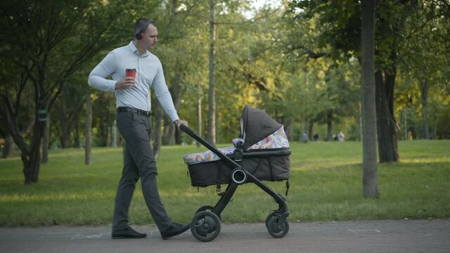 Wide shot of joyful young father in headphones walking with baby stroller along the alley in summer park, and singing. Side view portrait of cheerful Caucasian man enjoying walk with baby carriage.