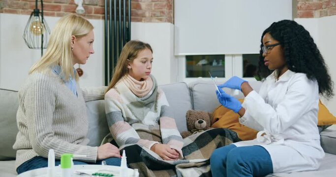 Attractive positive high-skilled black-skinned private female doctor giving an injection to unhealthy teen girl which coucht cold and recieves treatment at home under mother's supervision